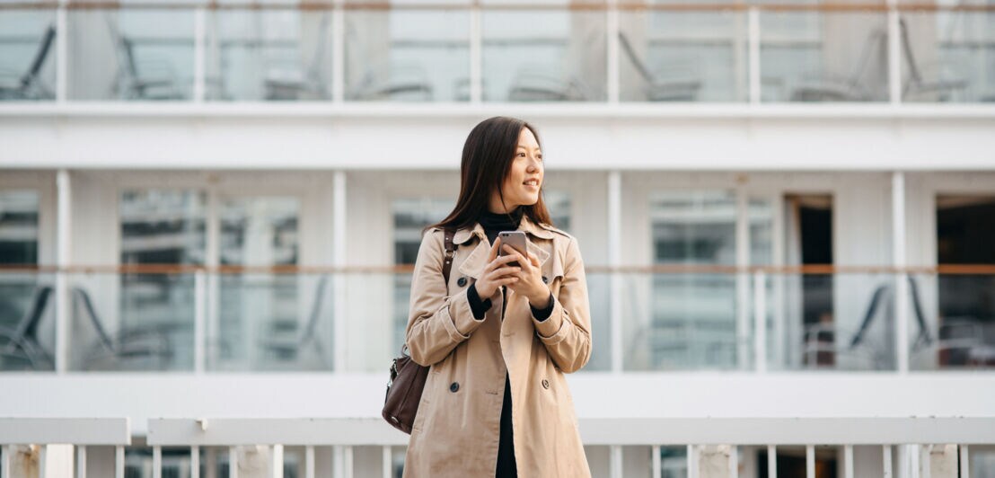 Eine Frau im Mantel steht mit ihrem Smartphone in den Händen vor einem Kreuzfahrtschiff.