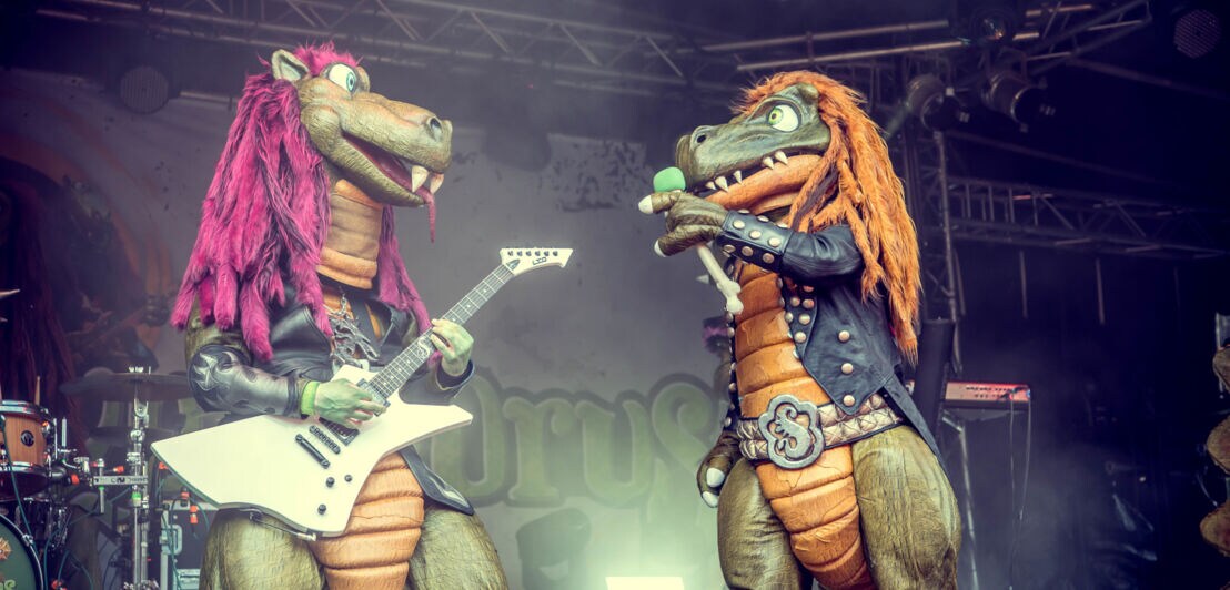 Eine Person im Drachenkostüm, die eine Gitarre hält und eine Person im Dinosaurierkostüm mit Mikrofon ansieht