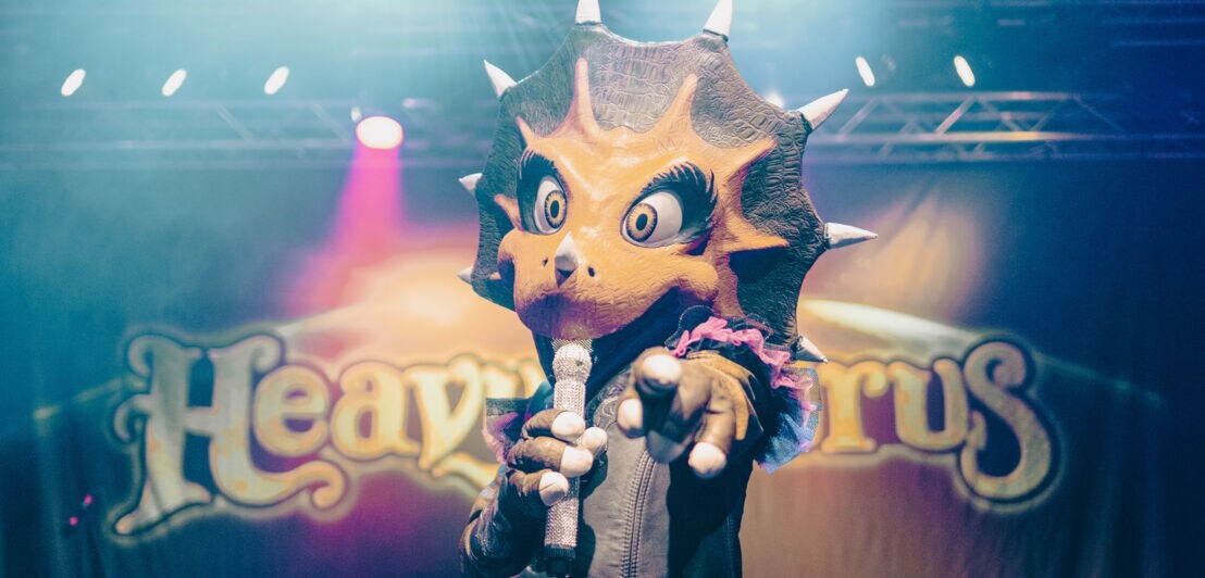 ine Person im Dinosaurierkostüm, die ein Mikrofon hält und mit einer Hand auf die Kamera zeigt, im Hintergrund Bühnenlicht und der Schriftzug „Heavysaurus“