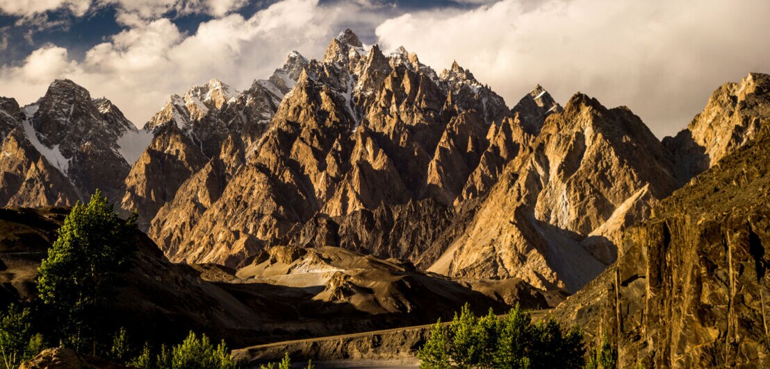 Sonnig beleuchtete, schroffe Bergspitzen mit Nadelbäumen im Vordergrund und bewölktem Himmel.