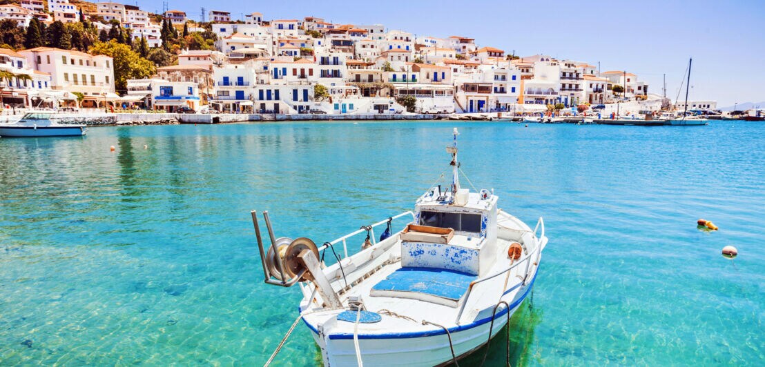 Ein Ort mit weißen Häusern im griechischen Stil an einer Meeresbucht, im Vordergrund ein Fischerboot im türkisblauen Wasser.