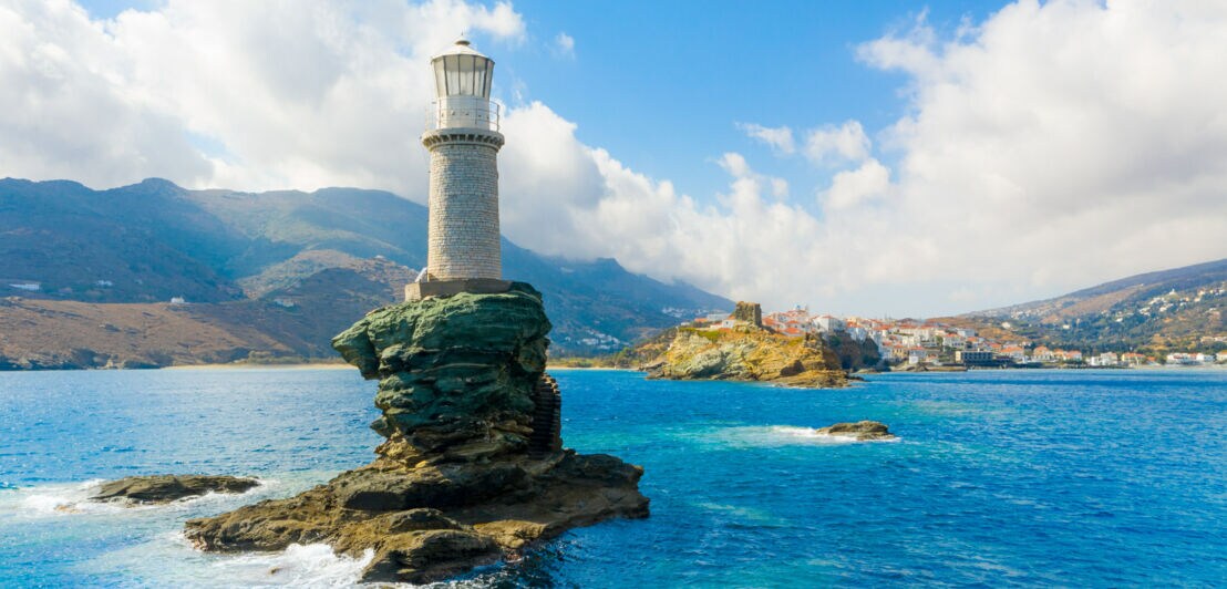 Weißer Leuchtturm auf einem kleinen Felsen im Meer vor einer Küste.