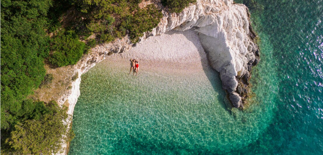Luftaufnahme eines Strandes mit zwei Personen, umgeben von grünen Bäumen und klarem türkisfarbenem Wasser