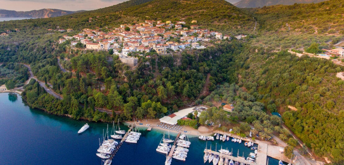 Luftaufnahme des Küstendorfes Meganisi mit Yachthafen, umgeben von bewaldeten Hügeln und ruhigem blauem Wasser