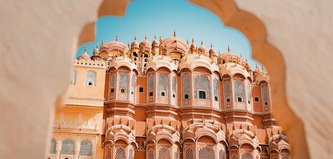Blick durch ein kunstvoll geformtes Tor auf den Hawa Mahal, ein rosa Sandsteinpalast mit vielen Fenstern in Jaipur