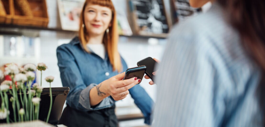 Kontaktlose Zahlung per Smartphone an einem Kartenlesegerät, gehalten von einer Frau hinter einer Theke in einem Geschäft.