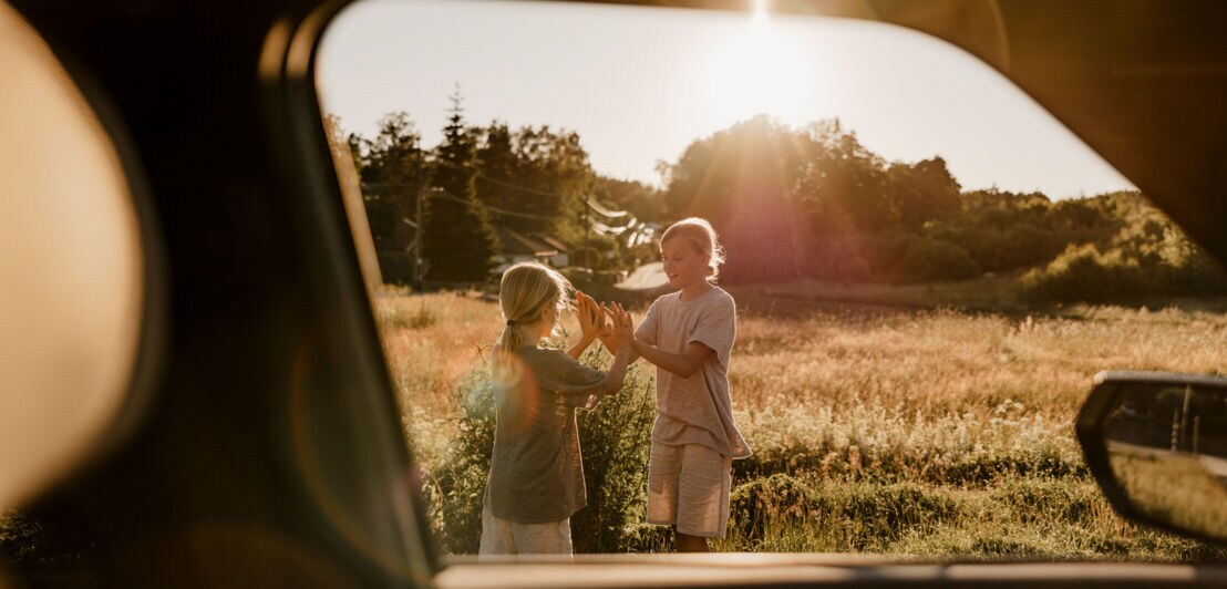 Blick durch ein geöffnetes Autofenster auf zwei spielende Mädchen auf einer Wiese im Sonnenschein.