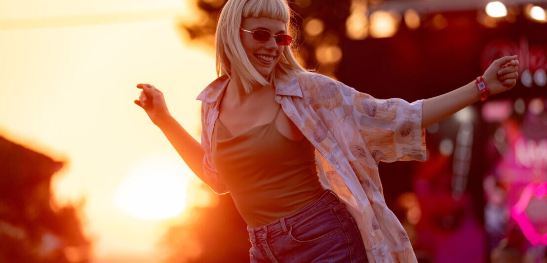 Eine Frau mit Sonnenbrille und in einem sommerlichen Outfit tanzt im Sonnenuntergang.