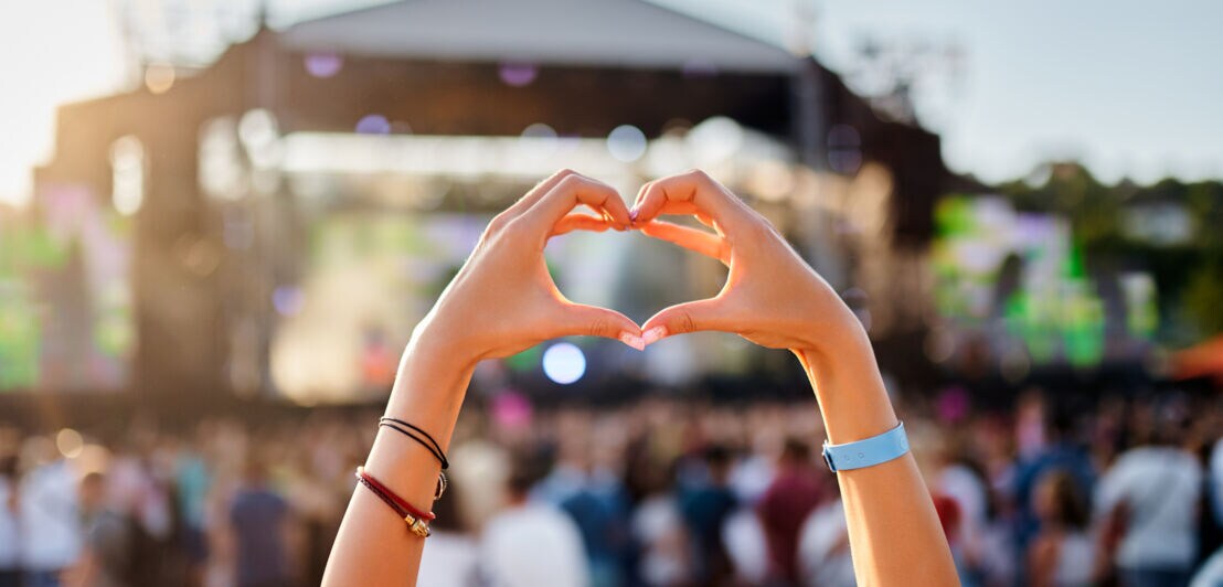 Die Hände einer Person formen ein Herz. Im Hintergrund ist eine Bühne zu sehen.