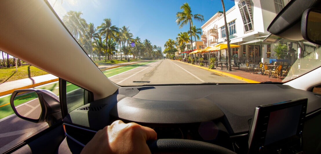 Aufnahme vom Fahrersitz eines Autos mit einer Hand am Lenkrad, das auf einer palmengesäumten Straße fährt