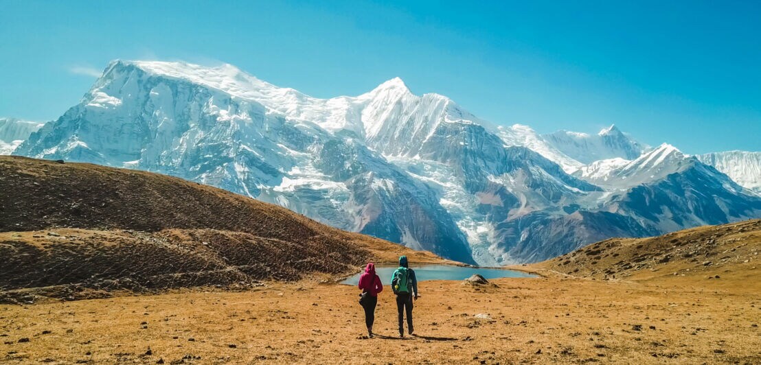 Zwei Personen auf einer Hochebene vor schneebedeckten Gipfeln des Himalayas.