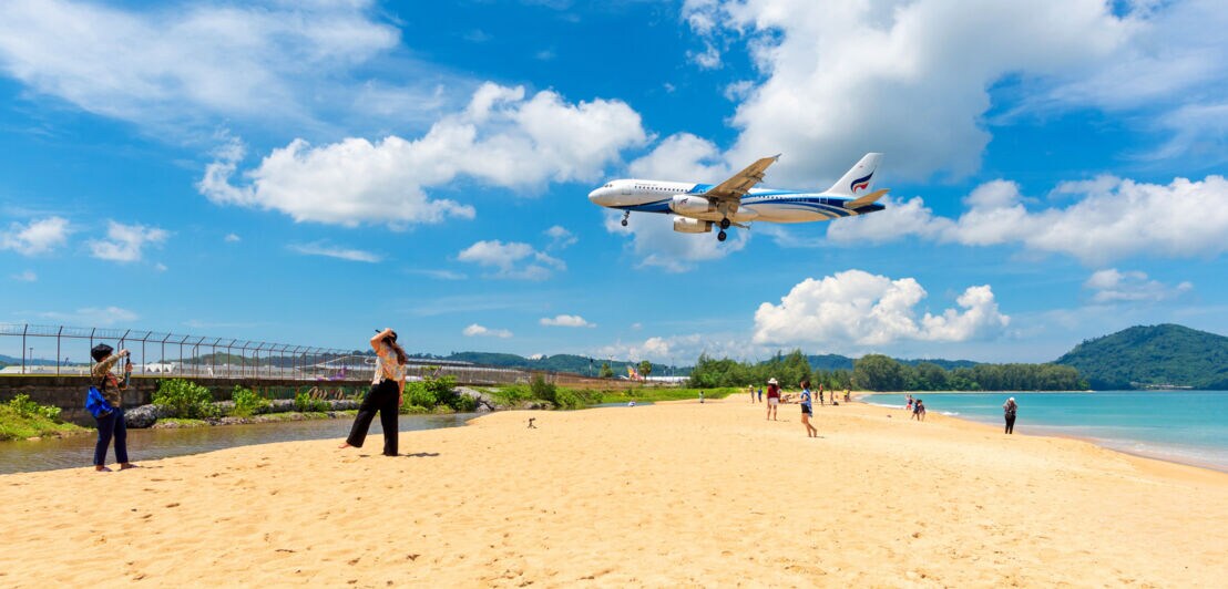 Personen auf einem Sandstrand an einem eingezäunten Gebiet, über dem ein Flugzeug im Landeanflug fliegt.