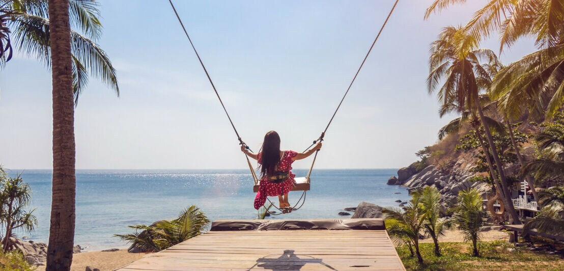 Rückansicht einer Frau im roten Kleid auf einer Schaukel an einem Strand, umgeben von Palmen.