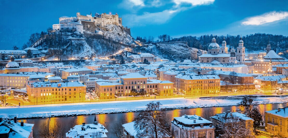 Schneebedecktes Stadtpanorama von Salzburg mit beleuchteten Straßen bei Dunkelheit.