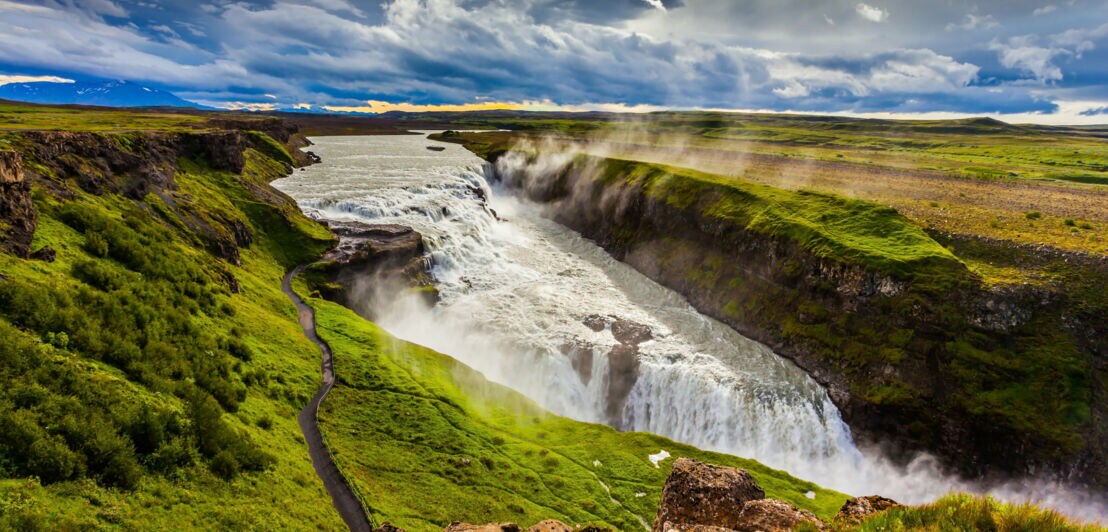 Breiter Wasserfall stürzt in zwei Stufen in eine tiefe Schlucht, umgeben von grünen Wiesen und bewölktem Himmel