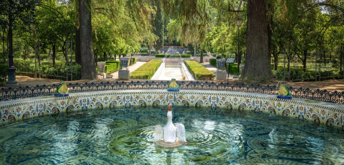 Verzierter Brunnen mit weißer Schwan-Skulptur in der Mitte, umgeben von Froschfiguren in einem Park mit Bäumen