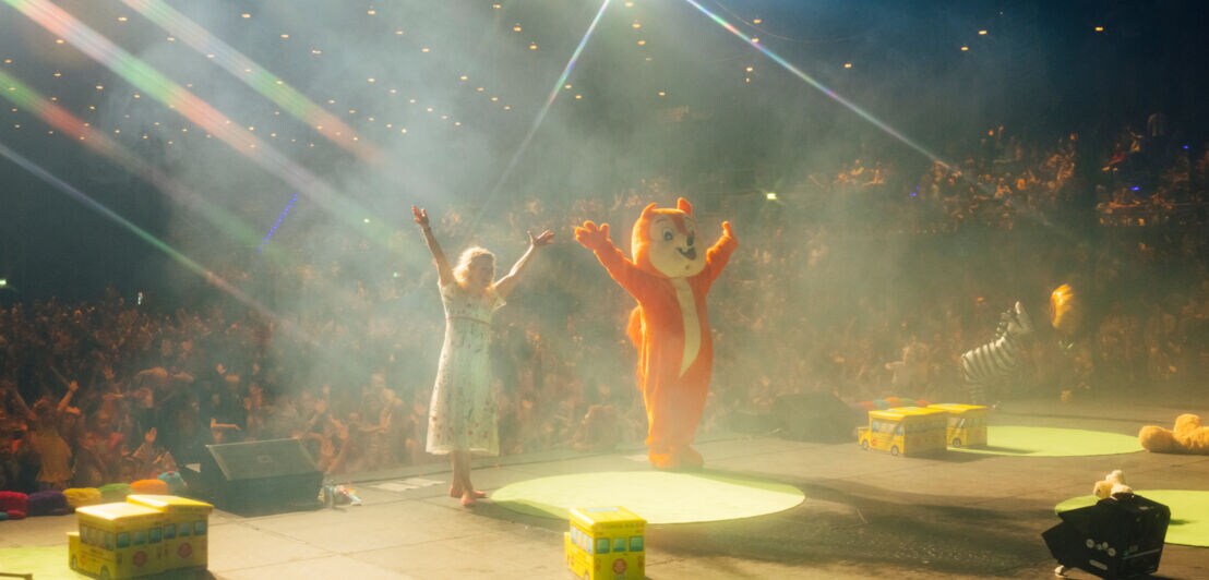 Simone Sommerland und eine Person im Eichhörnchen-Kostüm stehen mit weit ausgebreiteten Armen auf der Bühne mit dem Rücken zum Publikum.