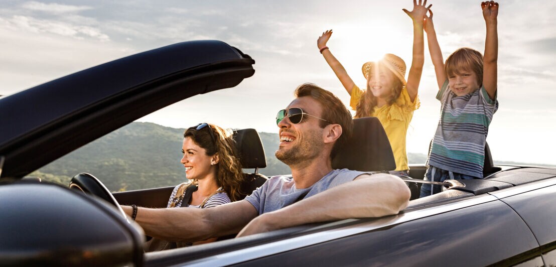 Eine Frau und ein Mann in einem offenen Cabriolet, auf den Rücksitzen zwei Kinder mit hochgestreckten Armen im Sonnenschein.