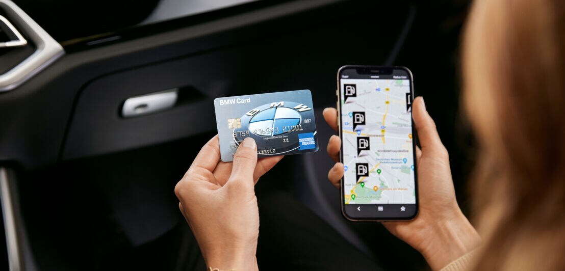 Eine BMW Card von American Express neben einem Smartphone mit Tankstellen-Icons auf einer Straßenkarte auf dem Display, gehalten von einer Person in einem Auto.