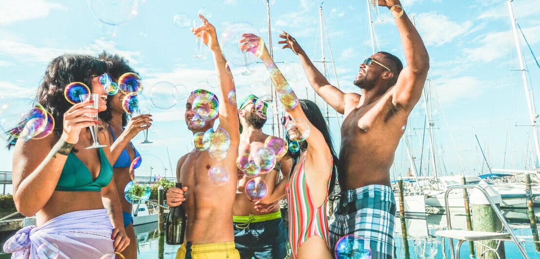 Freunde feiern und tanzen in Badebekleidung auf einem Boot, im Vordergrund sind Seifenblasen zu sehen.