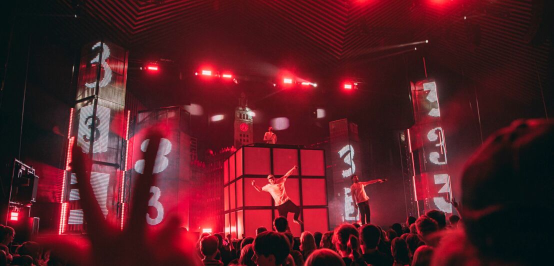 Musiker auf Bühne mit roter Beleuchtung und großen Bildschirmen, die die Zahl 3 zeigen, davor ist Publikum zu sehen.