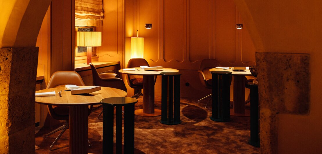 Stilvoll eingerichteter Gastraum mit runden Holztischen auf Teppichboden bei gedimmter, orangefarbener Beleuchtung.