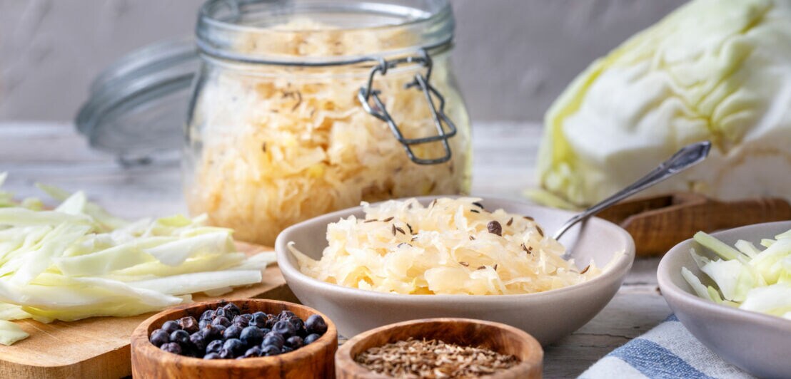 Schüssel mit Sauerkraut, Glas mit Sauerkraut, frischer Weißkohl, Schalen mit Wacholderbeeren und Kümmel auf Holztisch.