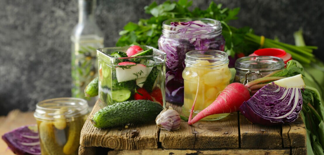 Verschiedene eingelegte Gemüse in Gläsern, darunter Gurken, Rotkohl und Zwiebeln, auf einem rustikalen Holztisch mit frischem Gemüse im Hintergrund.