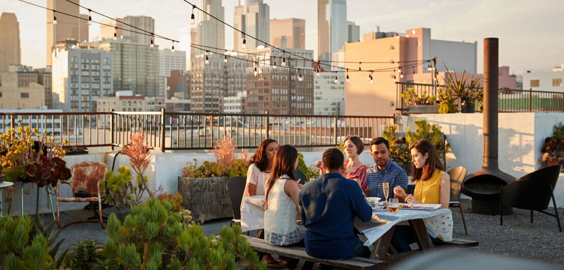 Sechs Personen sitzen beim Essen an einem Tisch auf einer Dachterrasse vor der Skyline einer Großstadt.