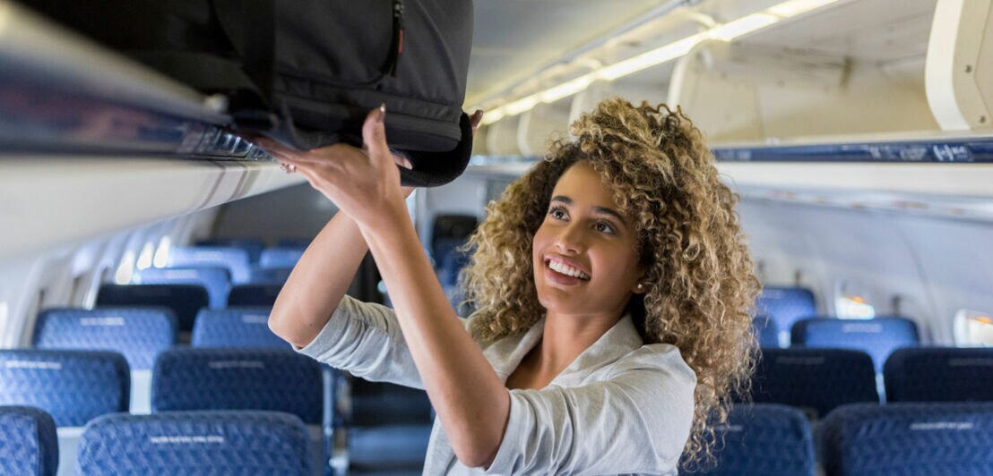 Eine junge Frau verstaut Handgepäck in einem Gepäckfach im Flugzeug.