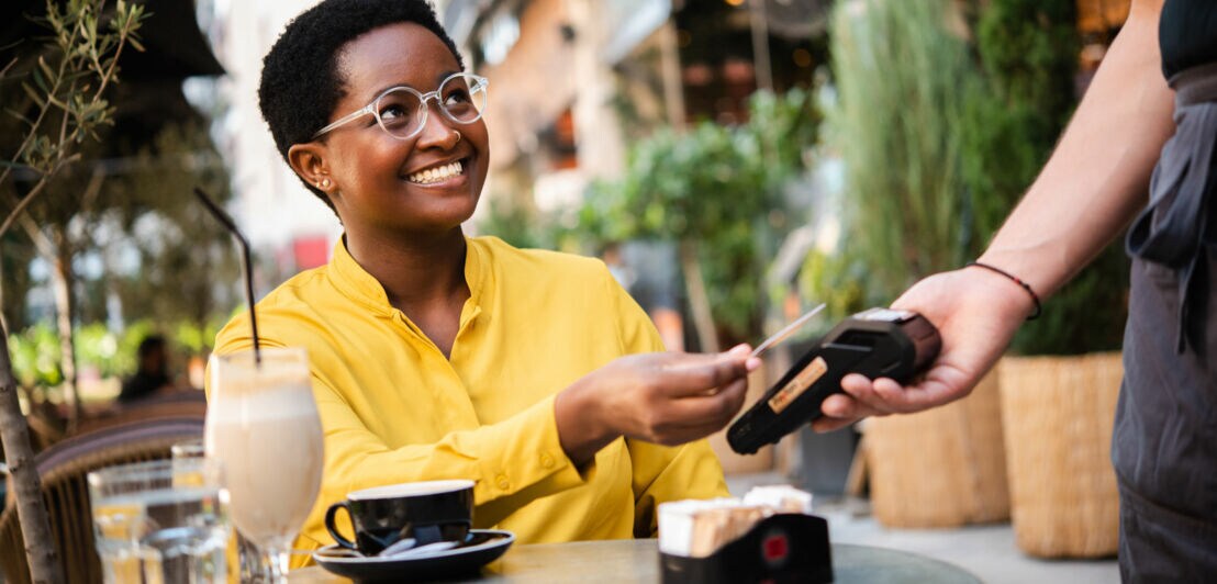 Eine lächelnde Frau bezahlt kontaktlos mit einer Kreditkarte an einem Kartenlesegerät an einem Tisch in der Außengastronomie.
