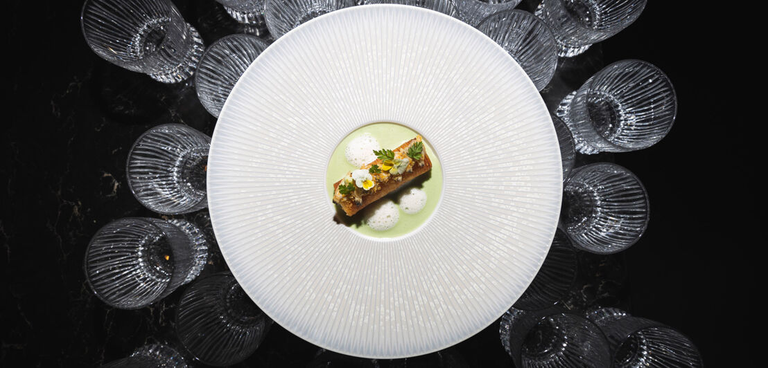 Aufsicht eines elegant angerichteten Gerichtes auf einem großen weißen Teller, der auf mehreren Kristallgläsern auf schwarzem Untergrund steht.