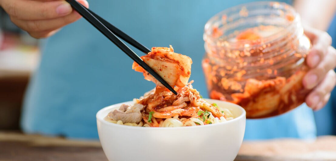 Eine Hand mit Essstäbchen platziert fermentierten Kohl auf einem Gericht in einer weißen Schale, dahinter ein Einmachglas mit Kimchi.