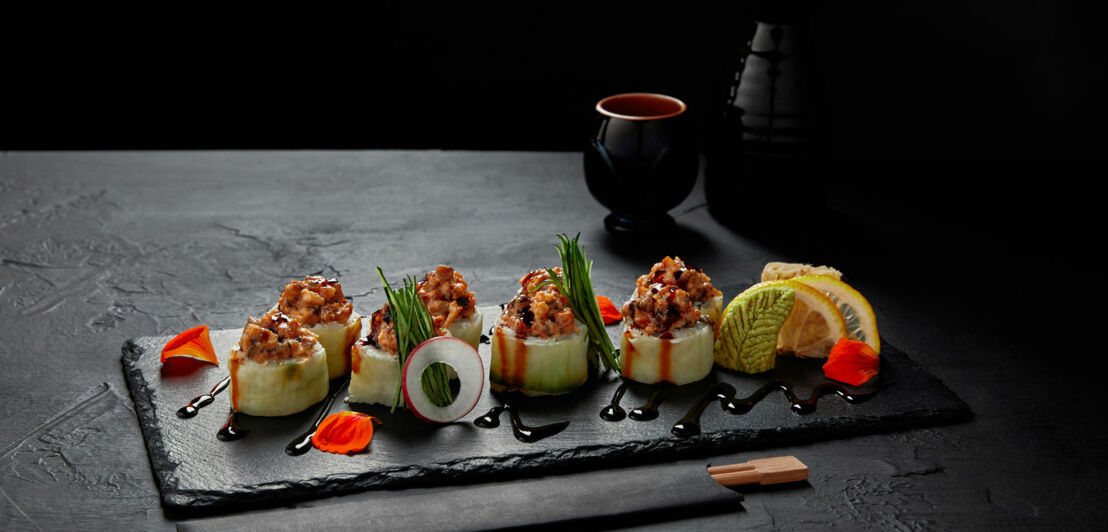 Sushi-Rollen mit rohem Fisch, Gurke, Frühlingszwiebeln und Sojasauce auf schwarzem Schiefer serviert, dekoriert mit Zitronenscheiben und Blütenblättern.