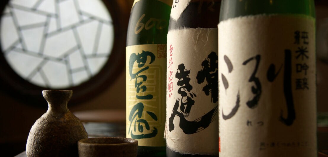 Drei dunkle Sake-Flaschen mit kunstvollen Etiketten neben einer kleinen Karaffe mit Becher.