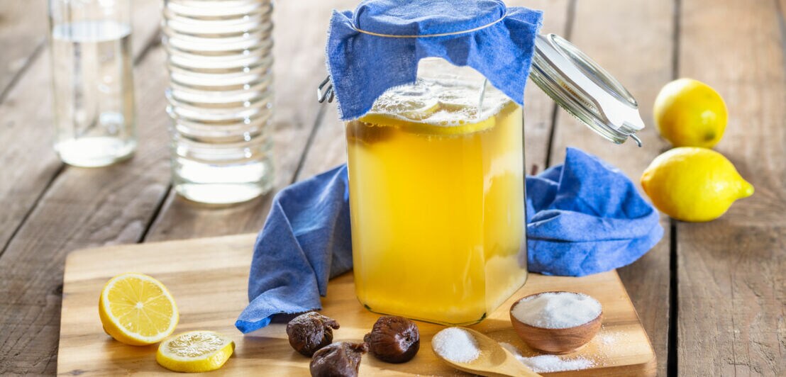 Ein Glasgefäß mit Wasserkefir bedeckt mit einem blauen Tuch, umgeben von Zitronen und weiteren Zutaten auf einem Holzbrett.