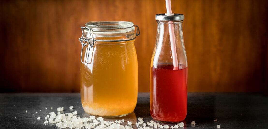 Ein Glas mit gelben und eine Flasche mit rotem Wasserkefir, umgeben von Kristallen auf dunklem Untergrund