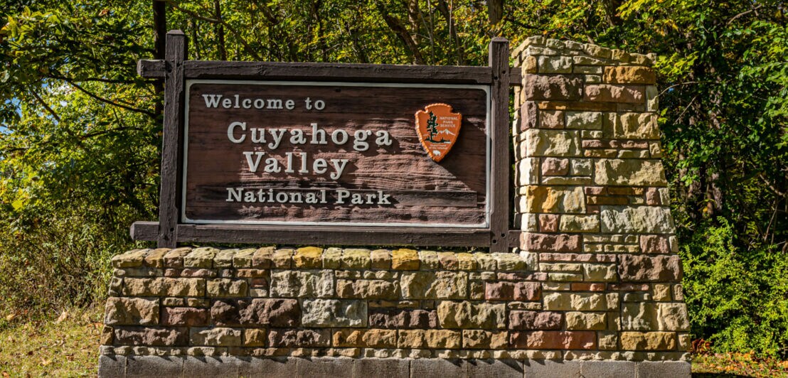 Willkommensschild des Cuyahoga Valley Nationalparks aus Holz auf einem steinernen Sockel, umgeben von Bäumen