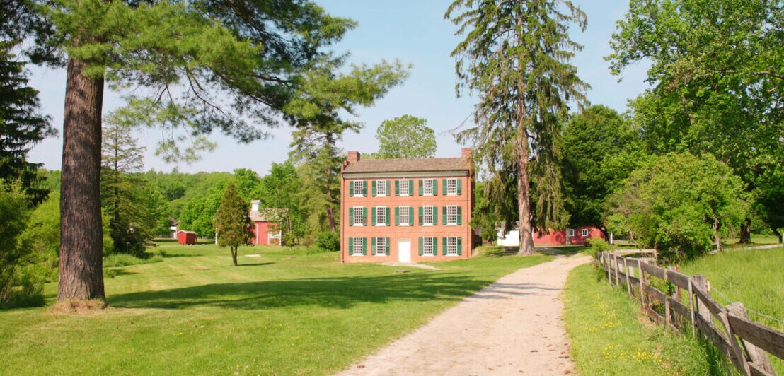 Backsteinhaus mit grünen Fensterläden am Ende eines sandigen Wegs, umgeben von großen Bäumen und Wiesen