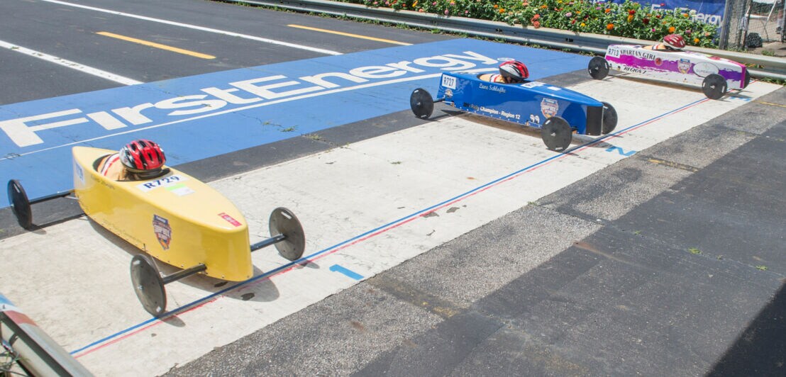 Drei seifenkistenartige Rennwagen in Gelb, Blau und Weiß an der Startlinie eines Rennens auf einer Straße 