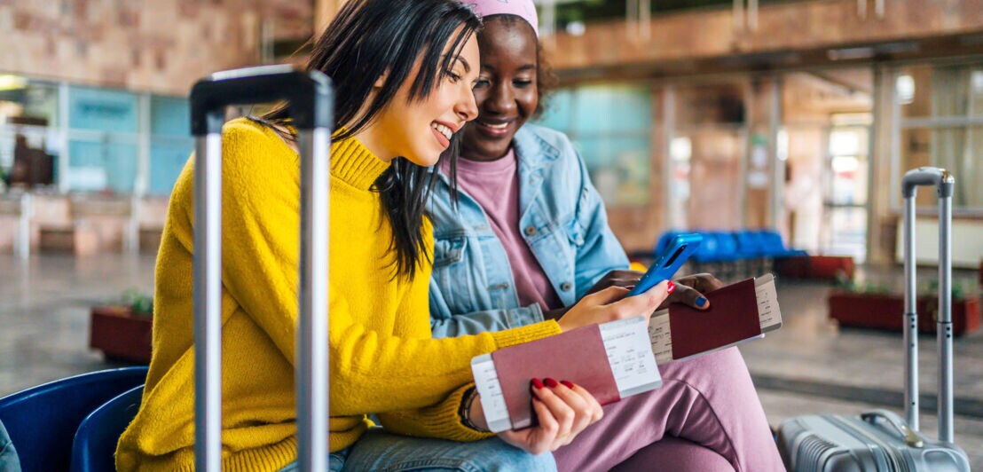 Zwei junge Frauen sitzen mit Trolleys, Reisepässen und Bordkarten in einem Terminal und schauen gemeinsam auf ein Smartphone.