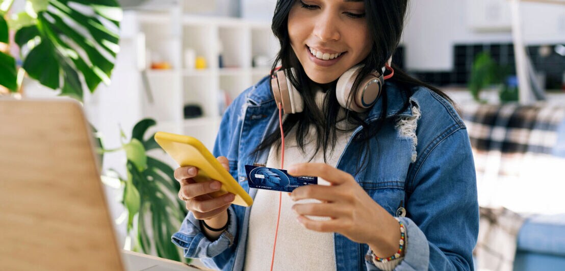 Eine junge Frau mit Kopfhörern sitzt mit einem Smartphone und einer blauen American Express Kreditkarte in den Händen vor einem Laptop am Schreibtisch in einer Wohnung.