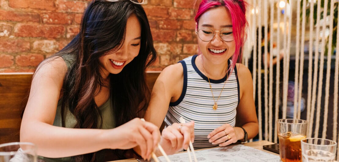 Zwei junge asiatische Frauen an einem Tisch in einem Restaurant greifen mit Essstäbchen nach einer Speise.