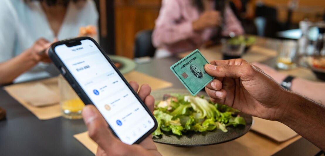 Eine grüne American Express Kreditkarte und ein Smartphone mit Amex App auf dem Display in den Händen einer Person an einem Tisch im Restaurant.