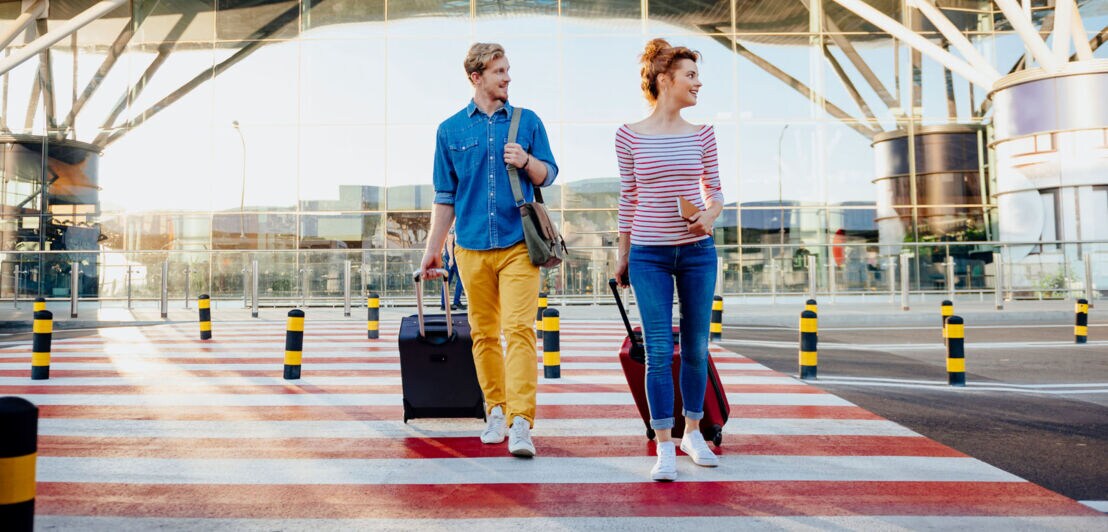Ein Mann und eine Frau laufen mit Rollkoffern über einen Zebrastreifen vor einem Flughafenterminal.