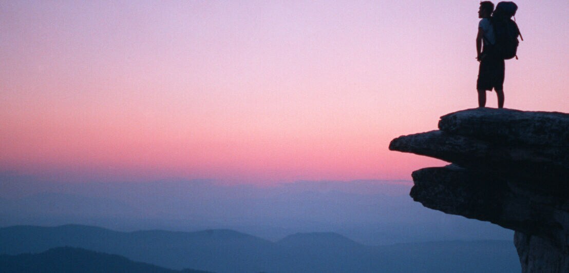 Wanderer mit großem Rucksack steht auf einem Felsen und blickt bei Sonnenuntergang über neblige Berglandschaft