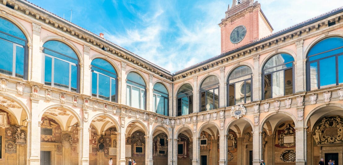 Innenhof eines historischen Gebäudes mit Arkadengängen, verzierten Säulen und einem Uhrturm unter blauem Himmel