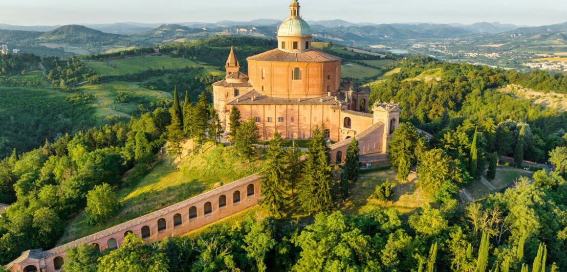 Historische Kuppelkirche auf einem bewaldeten Hügel mit umliegender Landschaft und Bergen im Hintergrund