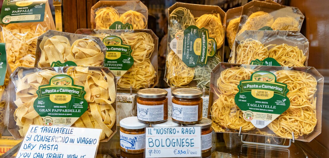 Verschiedene Packungen italienischer Pasta von La Pasta di Camerino und Gläser mit Bolognese-Sauce auf einem Regal, mit handgeschriebenen Preisschildern in Italienisch und Englisch