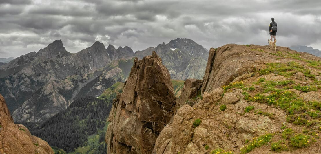 Wanderer steht auf einem felsigen Gipfel und blickt auf bewaldete Berge unter bewölktem Himmel